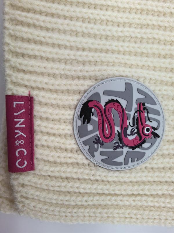 Bonnet hivernal en maille épaisse crème avec badge rond dragon rose et étiquette rose sur texture laine