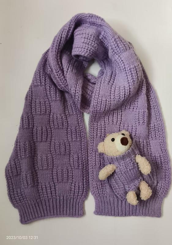 Écharpe tricotée violet lavande avec petit nounours crème dépassant d'une poche tricotée intégrée