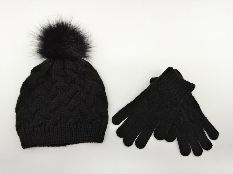 Bonnet câble noir pompom fourrure synthétique et gants tricotés assortis posés sur fond blanc