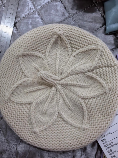 Béret tricoté crème avec relief motif floral feuilles torsadé au sommet posé à plat sur tissu argenté – béret femme chic hiver