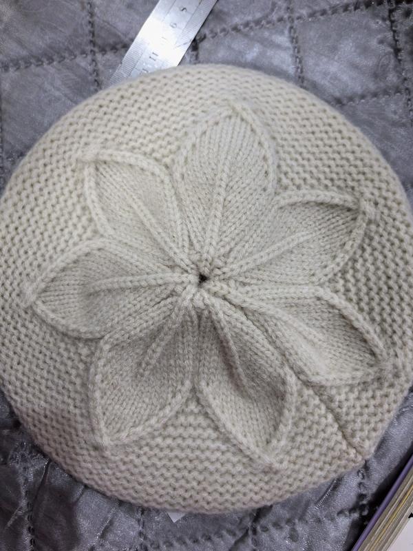 Vue de dessus béret tricoté crème avec motif fleur pétales torsadé en relief au sommet posé à plat sur tissu matelassé gris